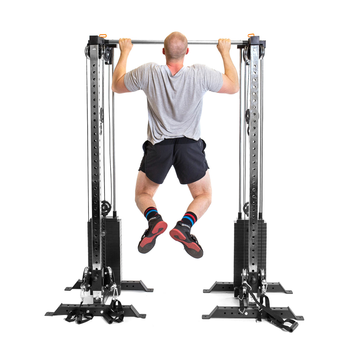 In their home gym, a person wearing a gray T-shirt and black shorts performs a pull-up on the Bells of Steel Cable Tower Squat Stands. Facing away from the camera, they expertly use the machine equipped with various weights and attachments.