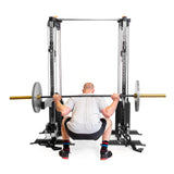 A person, dressed in a gray shirt, black shorts, multi-colored socks, and black shoes, performs a squat with a barbell on their shoulders using Bells of Steel's Cable Tower Squat Stands. The minimalist gym setting enhances the focus on their powerful posture as they face away.