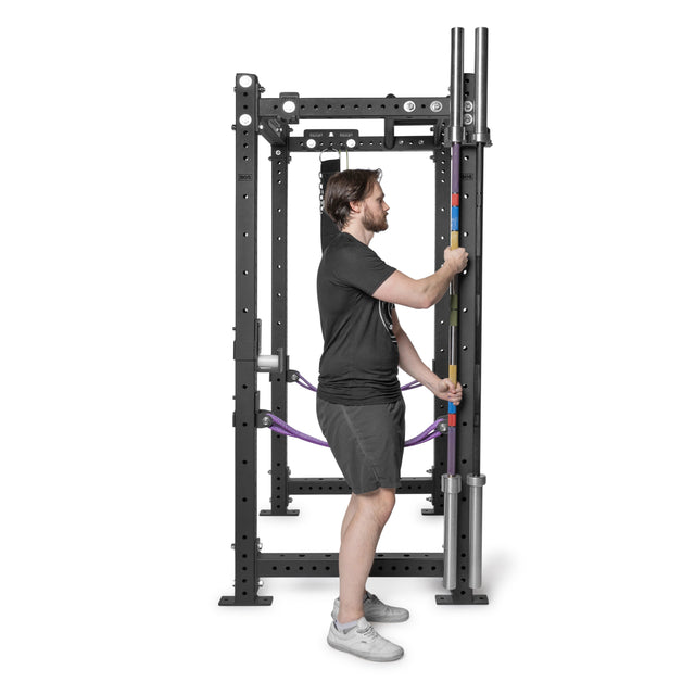 A man in a black t-shirt and shorts adjusts a colorful bar on a black power rack featuring Bells of Steel Hanging Barbell Holders in a gym.