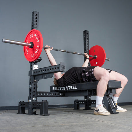 Wearing athletic gear, a person uses the Bells of Steel USA Manticore Two Post Converter Bench, lifting a barbell with red plates on a gym bench station.