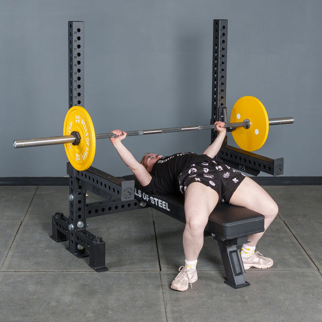 A person lies on the Manticore Two Post Converter Bench by Bells of Steel USA, gripping a barbell with yellow plates. The bench is set up by a gray wall and concrete floor, creating a classic gym station scene.