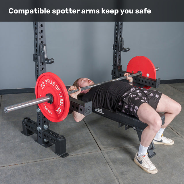 A person uses the Manticore Two Post Converter Bench by Bells of Steel USA, lying under a barbell with red plates in a power rack with spotter arms. Text above: "Compatible gym equipment spotter arms keep you safe.