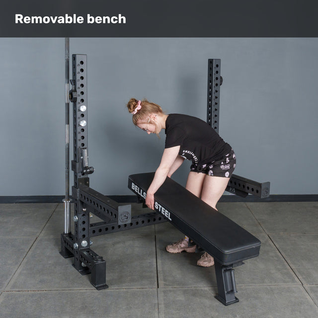 A woman adjusts a Manticore Two Post Converter Bench by Bells of Steel USA on a squat rack in a gym, demonstrating the bench station’s removability. "Removable bench" appears at the top, emphasizing versatile gym equipment.