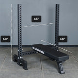 A Manticore Two Post Converter Bench by Bells of Steel USA sits under a 60-inch rack with a 43-inch horizontal bar. The 48-inch bench, placed on tiled flooring against a gray wall, creates an ideal bench press station.
