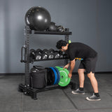 A man in athletic wear places a green weight plate onto the Bells of Steel USA Storage Shelves, which hold dumbbells, medicine balls, weight plates, a stability ball, and gym bags. The gym features gray walls and a tiled floor.
