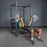 A person bench presses with a barbell inside a power rack, where Bells of Steel USA Storage Shelves hold various weight plates and kettlebells at the gym.