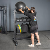 A man in athletic wear places a large exercise ball on the top Storage Shelves by Bells of Steel USA, which also hold weights, resistance bands, and other workout equipment.