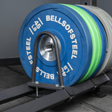 A close-up of Bells of Steel USA gym weight plates in blue and green stacked on a metal bar, with Storage Shelves and gym equipment visible on the floor.