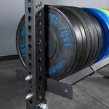 A close-up of Bells of Steel USA Storage Shelves holding several 45-pound black weight plates beside a power rack with numbered holes, set on a concrete floor against a gray wall.
