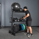 Wearing black workout clothes, a person organizes dumbbells on Bells of Steel USA Storage Shelves, which also hold medicine balls, resistance bands, bumper plates, and sandbags against a gray wall and floor.