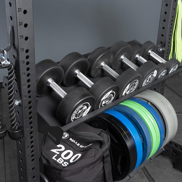 The Bells of Steel USA Storage Shelves hold five dumbbells, a 200-lb sandbag, colored weight plates, and have a rope attachment hanging on the side. The shelves are set against a gray wall and floor backdrop.