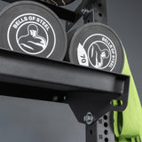 Two black "Bells of Steel USA" weight plates rest on Storage Shelves, with a green resistance band hanging nearby. The shelves and equipment stand out against the gray background.