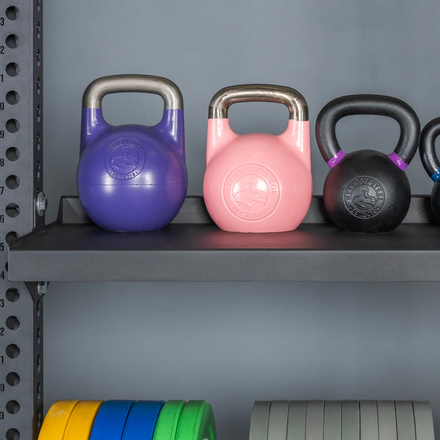 A Bells of Steel USA Storage Shelf holds purple, pink, and black kettlebells beside colorful weight plates on a lower shelf, all set against a gray gym wall.