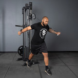 A man in a black t-shirt and shorts performs a cable crossover on the Bells of Steel USA Oblivyon Tower - 3" x 3” Cable Tower, standing with one foot forward and arms extended while holding the cable handles.