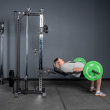 Wearing athletic gear, a person does a hip thrust with a barbell (green plates), upper back on a bench and feet flat beside the Bells of Steel USA Oblivyon Tower - 3" x 3" Cable Tower in the gym.
