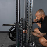 A bearded, tattooed man crouches down to adjust the weight pin on a Bells of Steel USA Oblivyon Tower - 3" x 3” Cable Tower in a plain, gray gym setting.