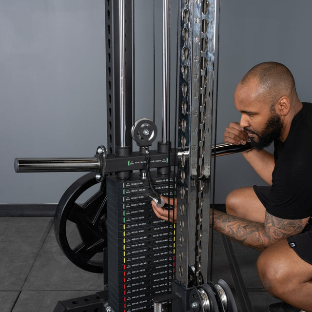 A bearded, tattooed man crouches down to adjust the weight pin on a Bells of Steel USA Oblivyon Tower - 3" x 3” Cable Tower in a plain, gray gym setting.