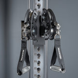 Close-up of the Bells of Steel USA Oblivyon Tower - 3" x 3” Cable Tower shows a black cable pulley system, adjustable numbered slots on uprights, and two "BELLS OF STEEL" handles hanging from carabiners in this home gym setup.