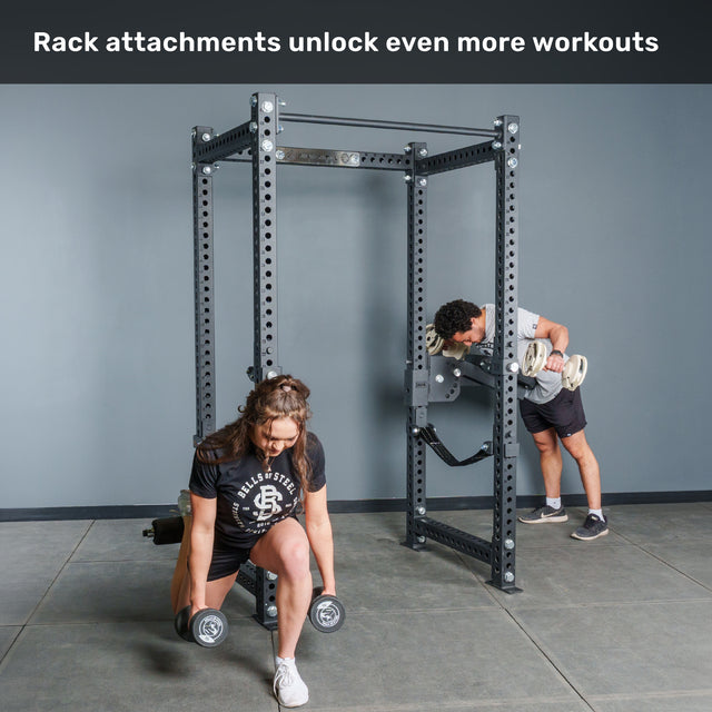 Two people work out in a gym with the Bells of Steel Manticore Four Post Power Rack Builder (3" x 3", 1" Holes). In front, a woman performs dumbbell lunges, while behind her, a man completes incline rows using attachments on the rack. Text above reads: "Rack attachments unlock even more workouts.