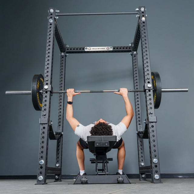 A person lies on a bench press, lifting a barbell loaded with weights inside the Bells of Steel Manticore Four Post Power Rack Builder (3" x 3", 1" Holes), viewed from the foot of the bench against a gray wall—an ideal configuration for any home gym power rack setup.