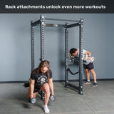 A woman does lunges with dumbbells while a man performs bicep curls using a Bells of Steel Manticore Four Post Power Rack - Prebuilt (3" x 3", 1" Holes) attachment in the gym. Text above reads, "Rack attachments unlock even more workouts.