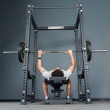 A person lies on a bench inside the sturdy Bells of Steel Manticore Four Post Power Rack - Prebuilt (3" x 3", 1" Holes), gripping a barbell with weights and preparing to perform a bench press exercise in their home gym.