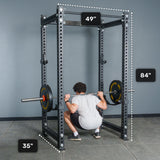 A person squats with a barbell inside the Bells of Steel Manticore Four Post Power Rack - Prebuilt (3" x 3", 1" Holes), finished in black and perfect for a home gym. The rack’s height is 84 inches, width is 49 inches, and depth is 35 inches, with each measurement clearly labeled around the equipment.