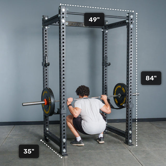 A person squats with a barbell inside the Bells of Steel Manticore Four Post Power Rack - Prebuilt (3" x 3", 1" Holes), finished in black and perfect for a home gym. The rack’s height is 84 inches, width is 49 inches, and depth is 35 inches, with each measurement clearly labeled around the equipment.