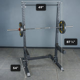 Standing tall against a gray wall on a tiled gym floor, the Bells of Steel Manticore Squat Stand - Prebuilt (3" x 3", 1" Holes) features sturdy black metal construction with substantial 3x3 uprights, paired with a barbell and weight plates. Measuring 87 1/4" in height, 49" in width, and 54" in depth, it makes an ideal centerpiece for any home gym.