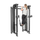 A person wearing a gray shirt and black pants is using Bells of Steel's Kraken Rack Attachment for Hydra & Manticore Racks to do pull-ups on a gray fitness power rack. This single-sided attachment includes pulleys and adjustable straps for versatile workouts.