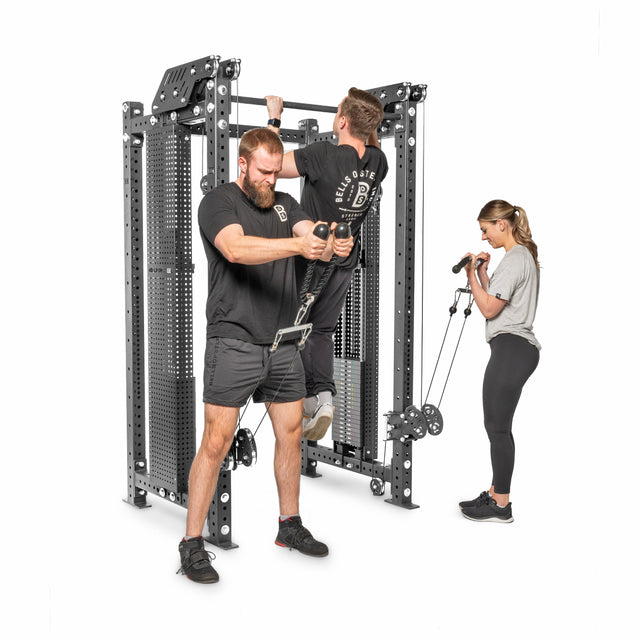 Three people use the Bells of Steel Kraken Rack Attachment for 4 Post Hydra & Manticore Racks. One does pull-ups, another uses the cable pulley for chest exercises, and a third performs bicep curls with resistance bands. The machine's adjustable trolleys keep everyone focused and engaged.