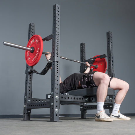 A person uses the Bells of Steel USA Four Post Converter Bench in a gym, lifting a barbell with red plates. They wear black shorts and shirt, white socks, and beige sneakers.