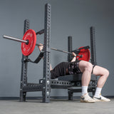A person performs a bench press with a barbell loaded with red plates on the Bells of Steel USA Hydra Four Post Converter Bench, shown from a low angle that highlights the bench, weights, and the lifter’s legs in a gym setting.