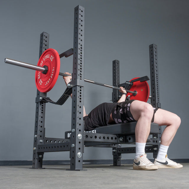 A person performs a bench press with a barbell loaded with red plates on the Bells of Steel USA Hydra Four Post Converter Bench, shown from a low angle that highlights the bench, weights, and the lifter’s legs in a gym setting.