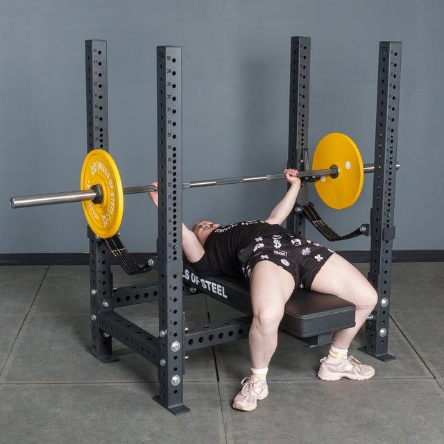 A person wearing black shorts and a light shirt is lying on the Hydra Four Post Converter Bench by Bells of Steel USA, gripping a barbell with yellow plates, preparing to lift in a gym with gray floor and wall.