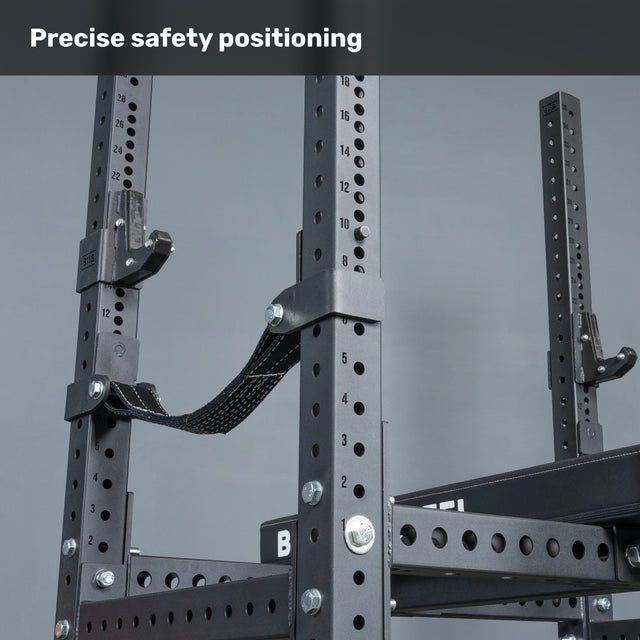 Close-up of the Bells of Steel USA Hydra Four Post Converter Bench, featuring black metal construction with numbered holes and attached safety straps for precise bench press safety. Plain gray background with text: "Precise safety positioning.