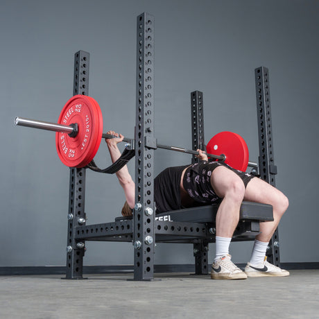 A person bench presses on the Manticore Four Post Converter Bench by Bells of Steel USA, featuring a floating black bench and red weight plates in a sturdy metal rack. The background is plain gray; the lifter wears shorts, T-shirt, and sneakers.
