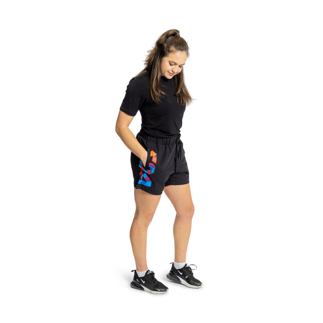 A woman stands on a white background wearing Bells of Steel black T-shirt and colorful patterned high-performance polyester Gym Shorts. Her hair is tied up as she looks down with hands in pockets, donning black athletic shoes crafted from stretchy fabric.