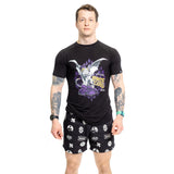 A man stands against a white background wearing a black and purple graphic T-shirt with "Outlaw King" and demon design. Tattooed arms complement his Bells of Steel Gym Shorts, black with white graphic designs, made of high-performance polyester for comfort. A digital watch is on his left wrist.