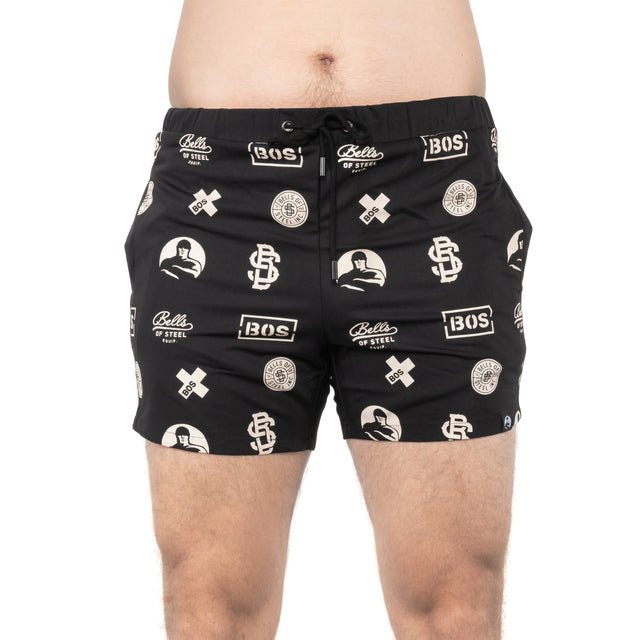 A person is wearing Bells of Steel black gym shorts featuring white logos and patterns like weights and "BOS" initials. Made from stretchy, high-performance polyester, they contour from waist to mid-thigh with a plain background.