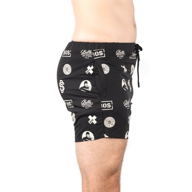 A model showcases Bells of Steel Gym Shorts in black with white logos and patterns. Made from high-performance polyester, the stretchy fabric is depicted in a side view extending from the waist to above the knee against a plain white background.