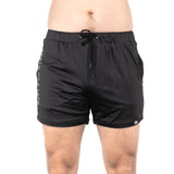 A person wearing Bells of Steel Gym Shorts, featuring a black color and a drawstring waistband, stands against a plain white background. These stretchy shorts in high-performance polyester fabric ensure comfort and flexibility, with "ISLE OF" partially visible on the side.