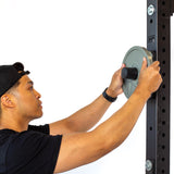A person in a black cap and t-shirt attaches Olympic plates to Bells of Steel Bolt-On Plate Pegs mounted on a black power rack, shown sideways against a white background.