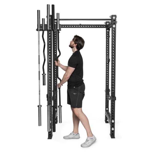 A man in black athletic wear stores a barbell using Bells of Steel Hanging Barbell Holders attached to a power cage, which provide convenient barbell storage.
