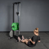A woman in black workout clothes and pink sneakers uses the Bells of Steel USA Footplate for Cable Tower/All In One Trainer to perform a seated row, with green and white weights nearby against a plain gray wall.
