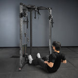 A person in black athletic wear sits on the floor before a Bells of Steel USA Footplate for Cable Tower/All In One Trainer, preparing to exercise in a gym with dark floors, dark walls, metallic equipment, and black accents.