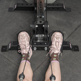 A person in pink sneakers and cartoon socks sits at a gym rowing machine, feet on the Bells of Steel USA Footplate for Cable Tower/All In One Trainer, gripping rower cables—a versatile accessory for effective workouts.