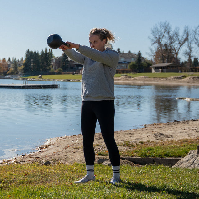 A woman exercises outdoors by a lake, swinging a Bells of Steel 6-12KG Adjustable Kettlebell with both hands. She wears a gray sweater, black leggings, and white socks. Trees and houses are visible in the background.