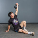 Wearing athletic gear, a person performs a Turkish get-up exercise on a gym floor, holding the Bells of Steel 6-12KG Adjustable Kettlebell overhead with one arm while supporting themselves with the other hand.