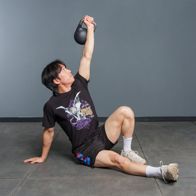 Wearing athletic gear, a person performs a Turkish get-up exercise on a gym floor, holding the Bells of Steel 6-12KG Adjustable Kettlebell overhead with one arm while supporting themselves with the other hand.